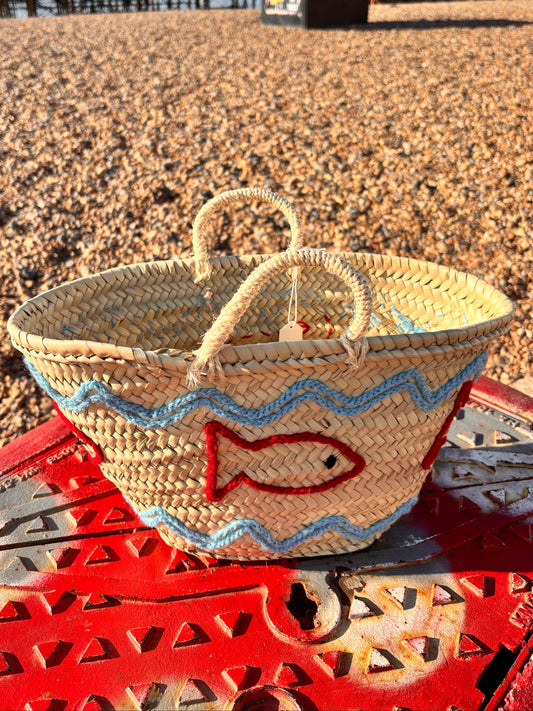 Decorative straw basket with red and blue embroidered fish and wave patterns. Photographed infront of the Brighton Palace Pier.