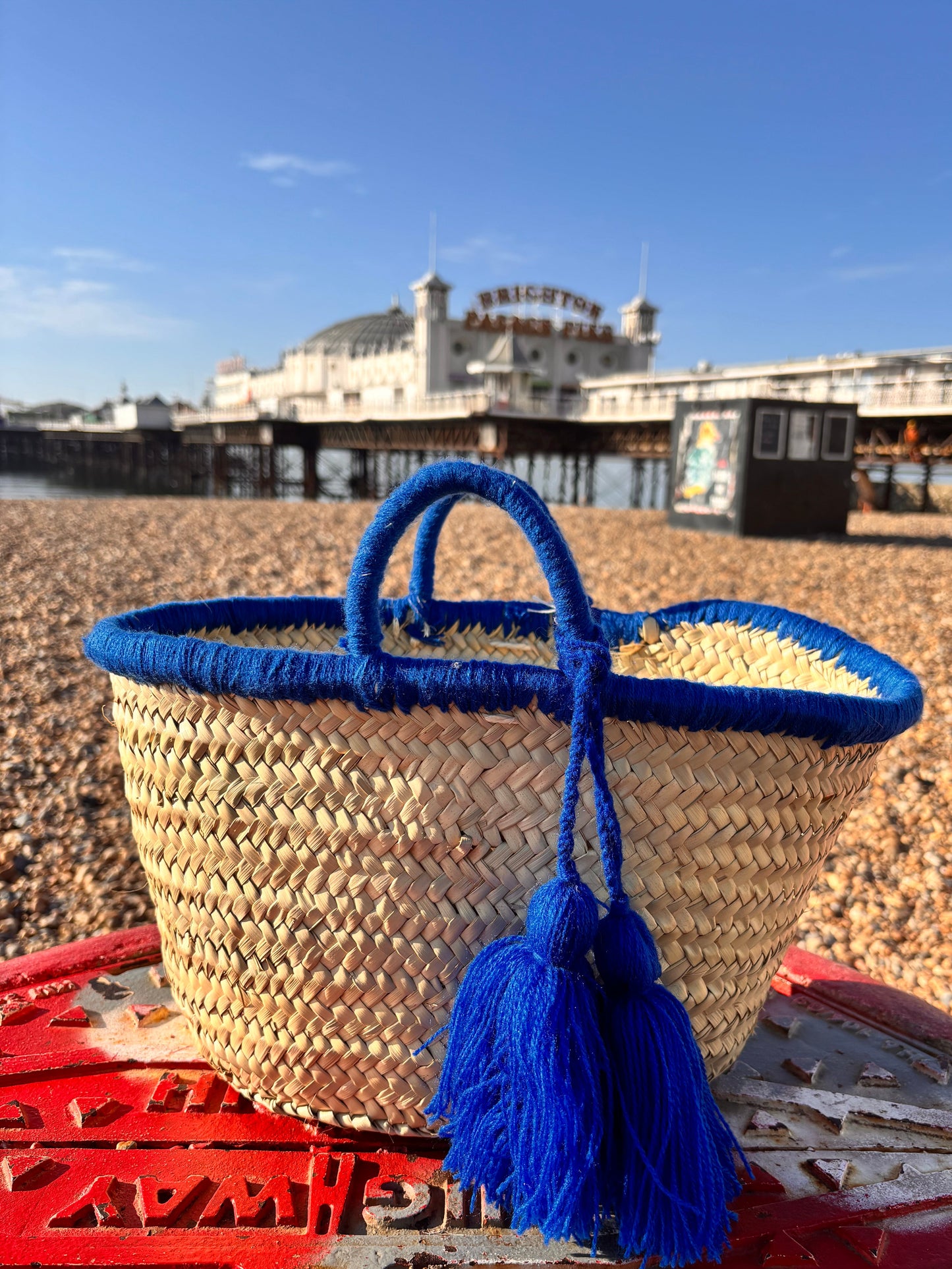 Handwoven Moroccan straw basket with bright blue wool tassels and blue-wrapped handles, placed on Brighton beach with Brighton Pier in the background under a clear blue sky.
