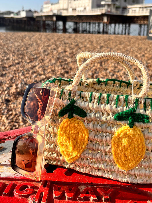 Small handwoven Moroccan straw bag with two crocheted yellow lemons and green embroidered leaves on the front. The bag is photographed on Brighton beach with Brighton Pier in the background, resting on a red painted metal surface alongside a pair of sunglasses.