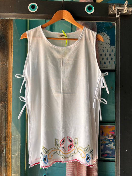 White sleeveless top with colorful embroidery on a hanger against a textured wall.