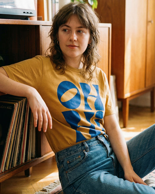 Person wearing a yellow 'Oh Hi' t-shirt with a blue retro text, sitting on the floor in a room with wooden furniture and plants.