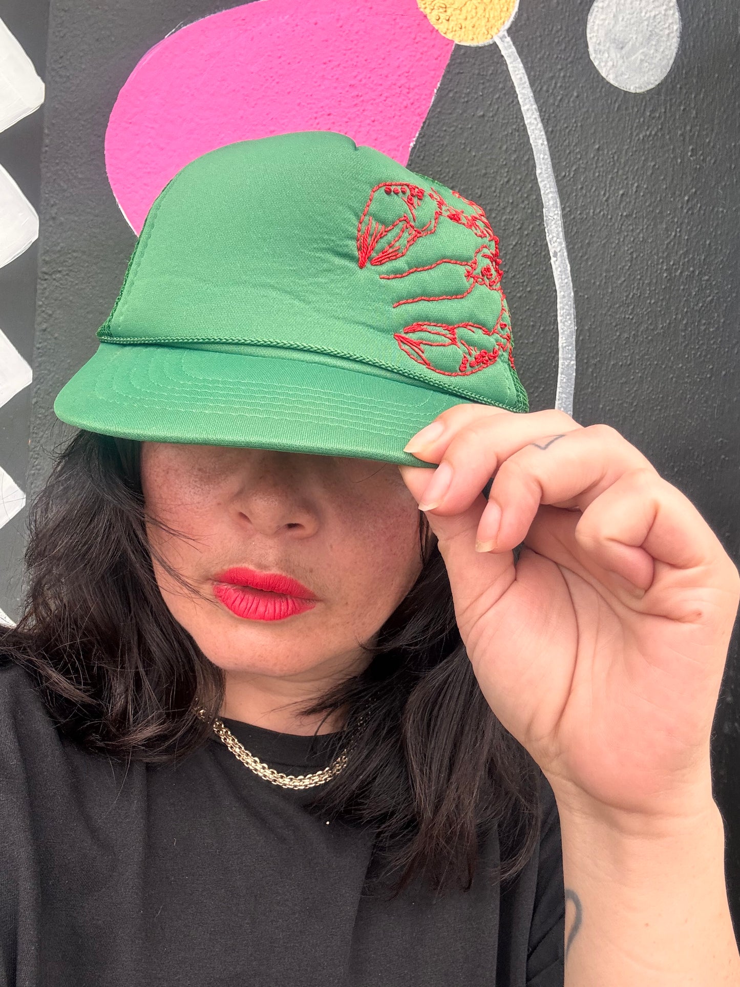Person holding a customised green trucker cap with red lobster embroidery against a colourful background