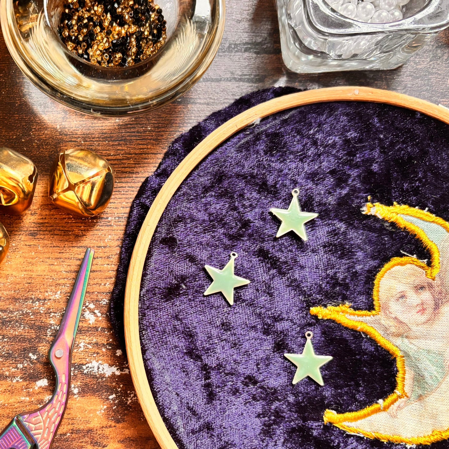Embroidery hoop with celestial design on purple fabric, surrounded by decorative items on a wooden surface.