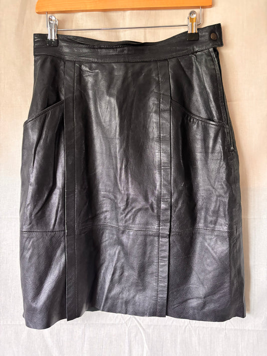 Front view of a Black leather skirt hanging on a wooden hanger against a beige wall.