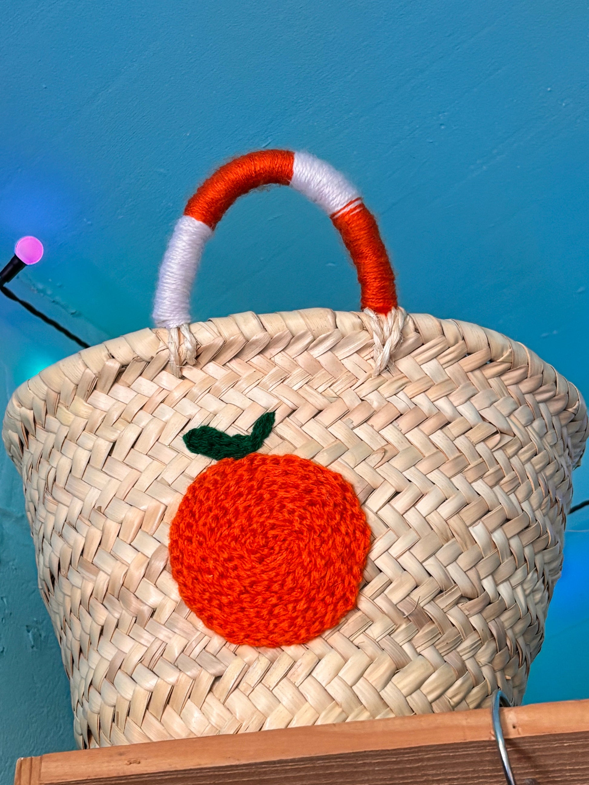 Handwoven Moroccan straw basket with a bright orange embroidered fruit detail and orange-and-white wrapped handles, displayed against a turquoise wall.