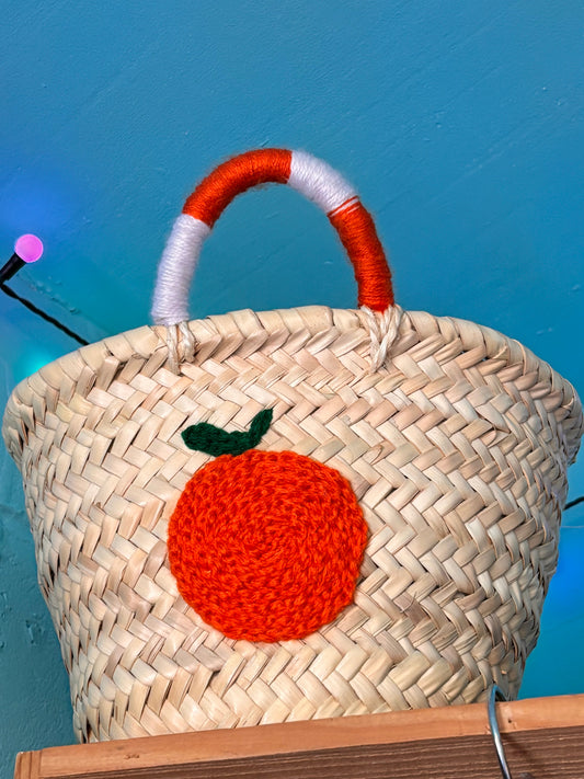 Handwoven Moroccan straw basket with a bright orange embroidered fruit detail and orange-and-white wrapped handles, displayed against a turquoise wall.