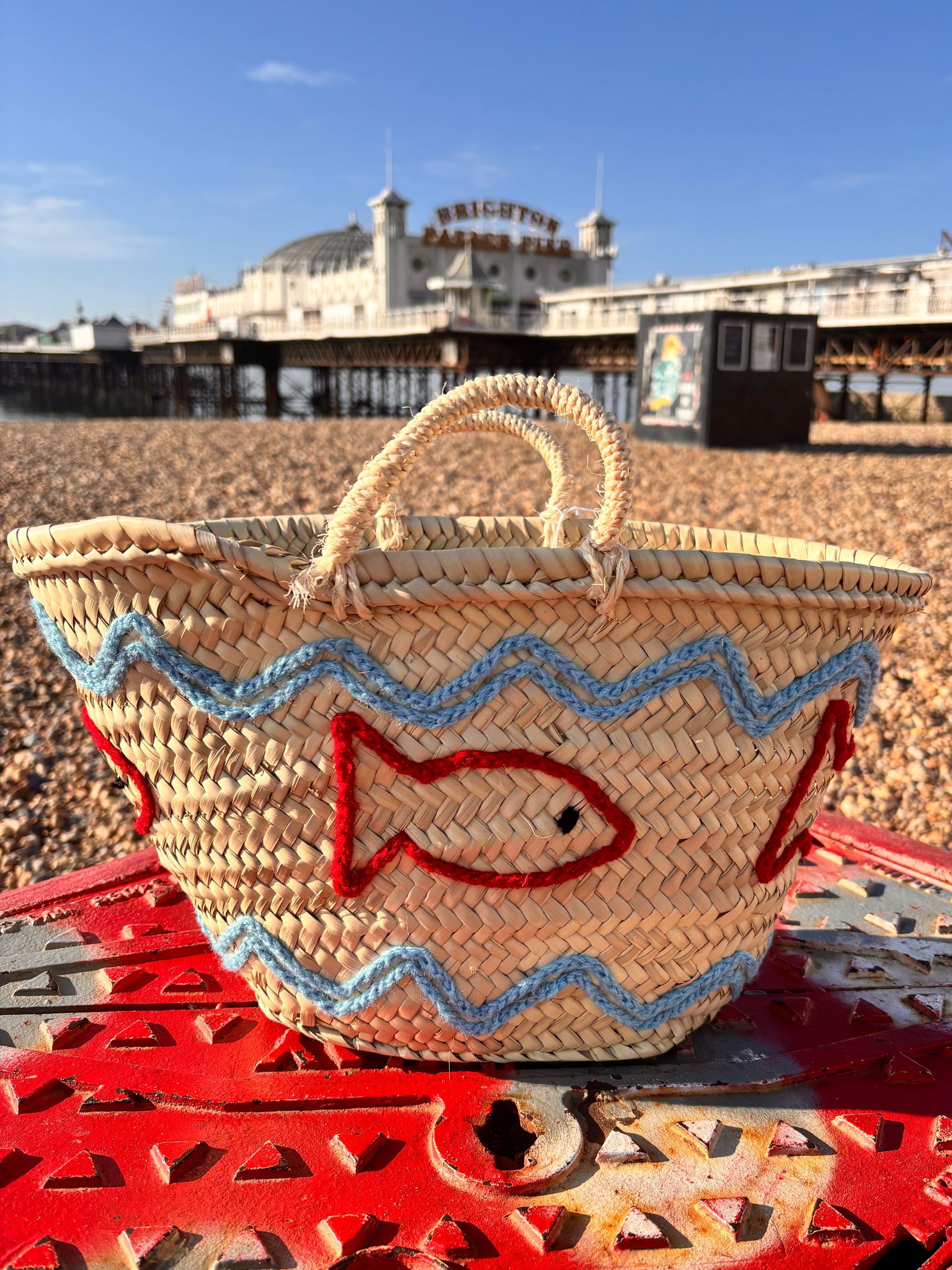 Decorative straw basket with red and blue embroidered fish and wave patterns. Photographed infront of the Brighton Palace Pier.
