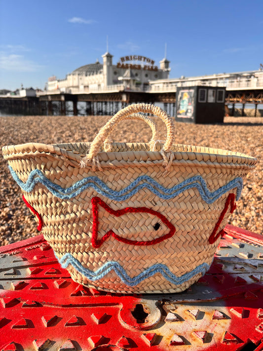 Decorative straw basket with red and blue embroidered fish and wave patterns. Photographed infront of the Brighton Palace Pier.
