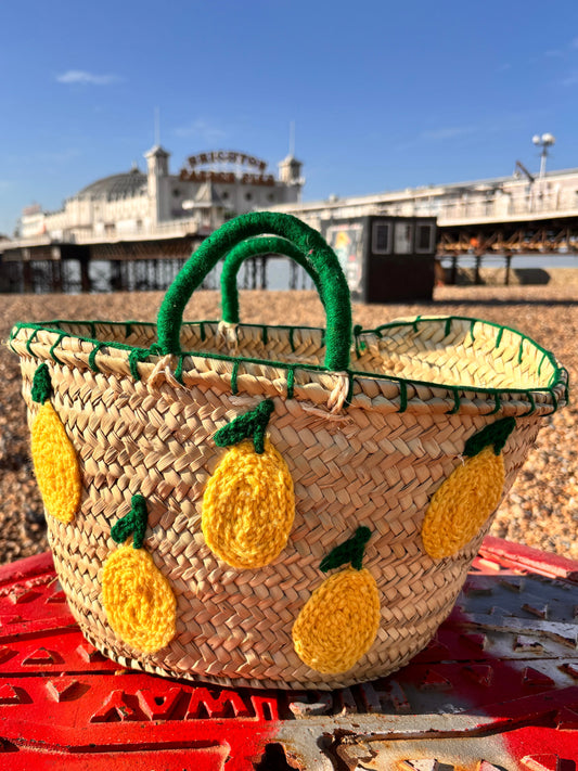 Handwoven Moroccan straw basket decorated with crochet yellow lemons and green leaves, featuring green wool-wrapped handles, photographed on Brighton beach with the pier in the background.