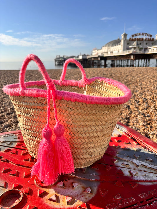 Handwoven Moroccan straw basket with bright pink wool tassels and pink-wrapped handles, placed on Brighton beach with Brighton Pier in the background under a clear blue sky.