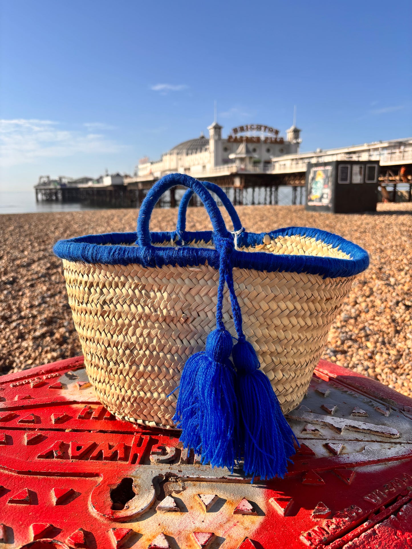 Handwoven Moroccan straw basket with bright blue wool tassels and blue-wrapped handles, placed on Brighton beach with Brighton Pier in the background under a clear blue sky.