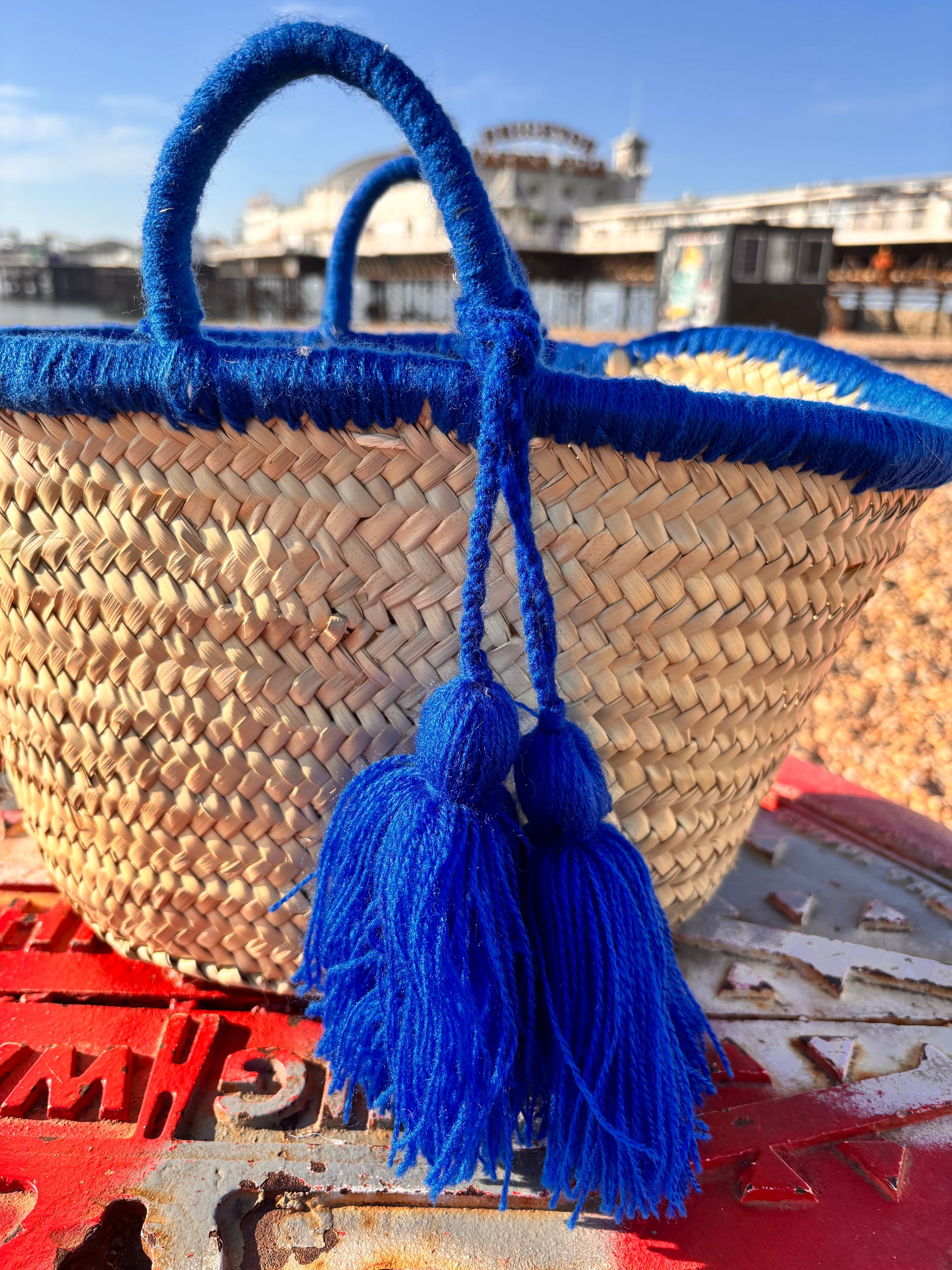 Handwoven Moroccan straw basket with bright blue wool tassels and blue-wrapped handles, placed on Brighton beach with Brighton Pier in the background under a clear blue sky.
