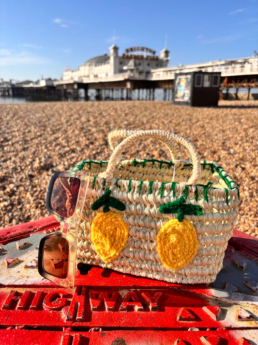 Small handwoven Moroccan straw bag with two crocheted yellow lemons and green embroidered leaves on the front. The bag is photographed on Brighton beach with Brighton Pier in the background, resting on a red painted metal surface alongside a pair of sunglasses.