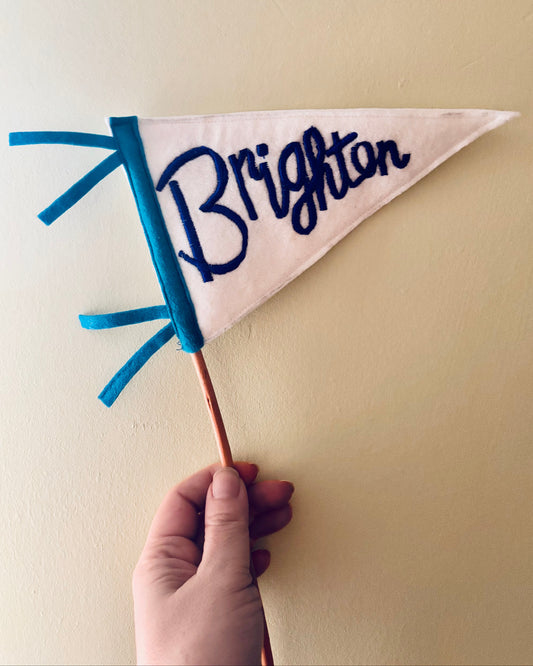 Hand holding a white pennant flag with blue trim and 'Brighton' embroidered in dark blue