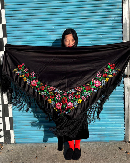 Person holding a black shawl with floral embroidery in front of a colorful wall.