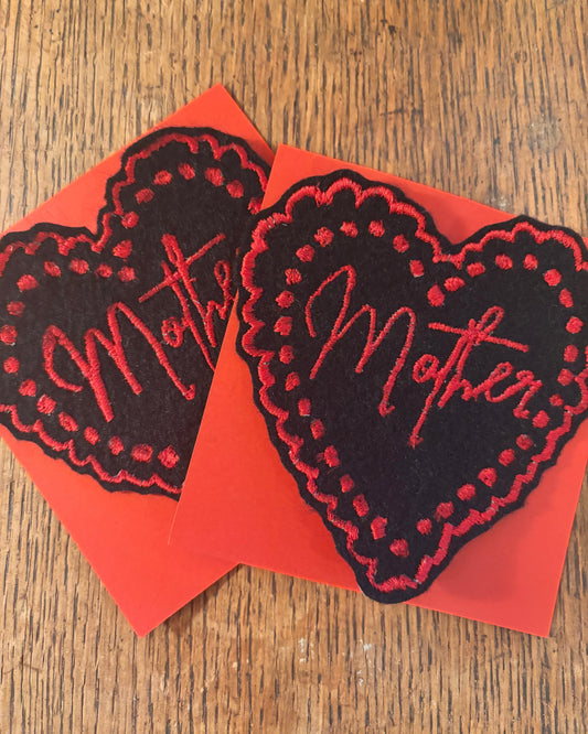 Two black heart-shaped embroidery patches with red detail and 'mother'. On a wooden surface.