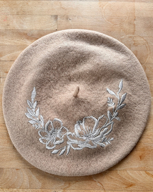 Beige beret with floral embroidery in light silver on a wooden surface