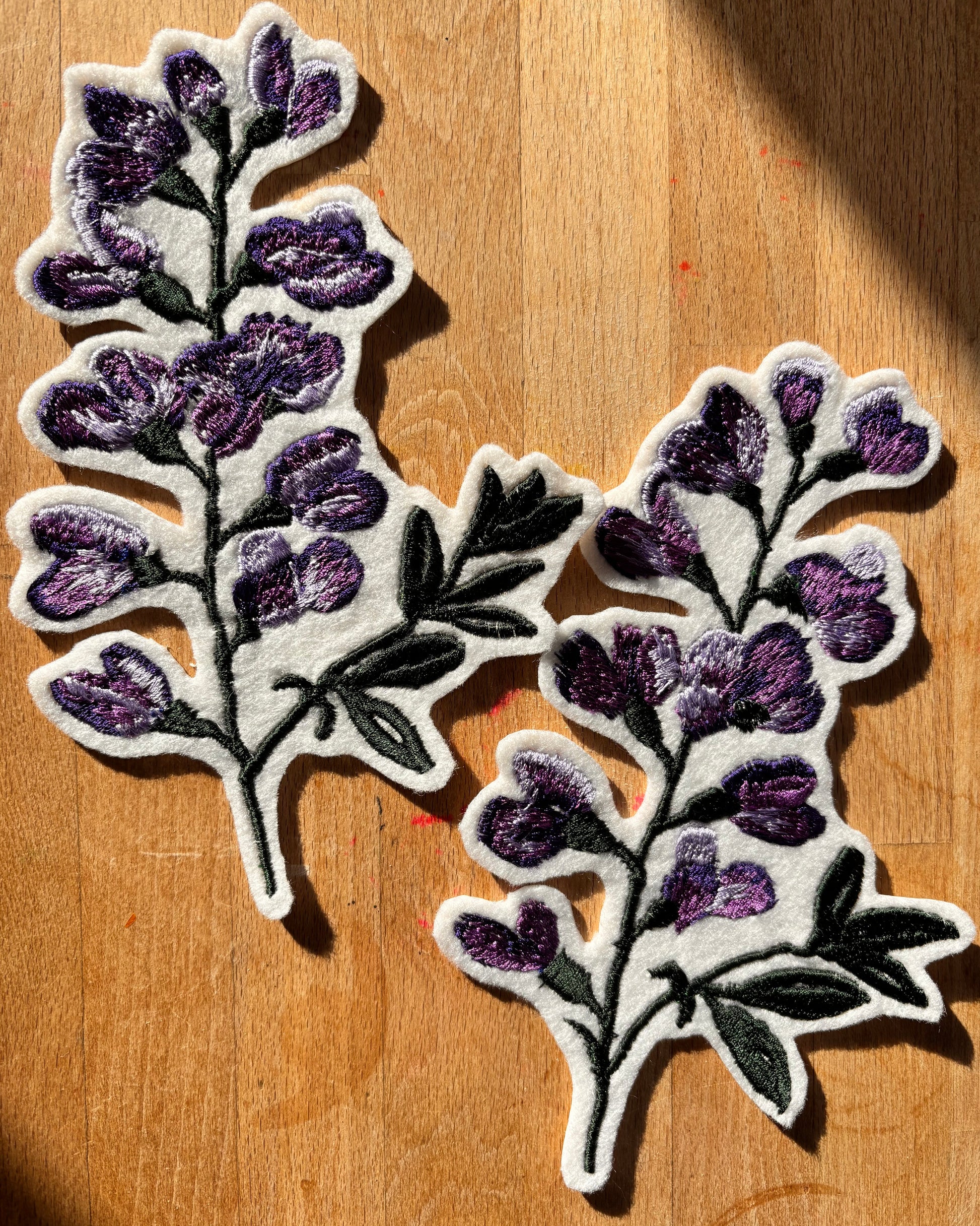 Two floral embroidery patches with purple flowers and green leaves on a wooden surface.