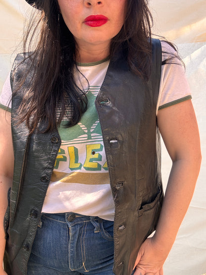 Person wearing a black leather vest over a white shirt with green text and blue jeans.