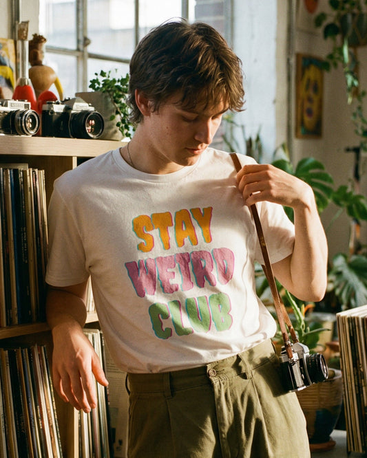 Person wearing a white 'Stay Weird Club' t-shirt with colourful text in a room filled with books and records.